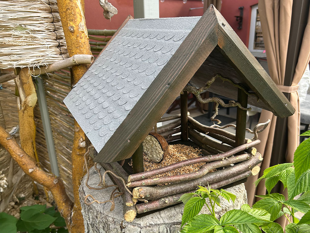 Selbstgebautes Vogelhaus aus Ästen und Holz mit grauem Dach, montiert auf einem Baumstumpf mit Vogelfutter im Inneren.