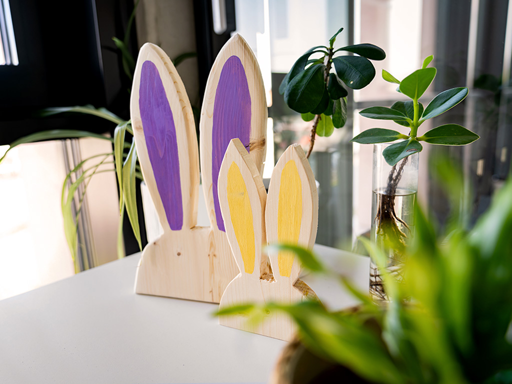 Zwei dekorative Holzhasen mit bemalten Ohren stehen auf einem Tisch neben Zimmerpflanzen.