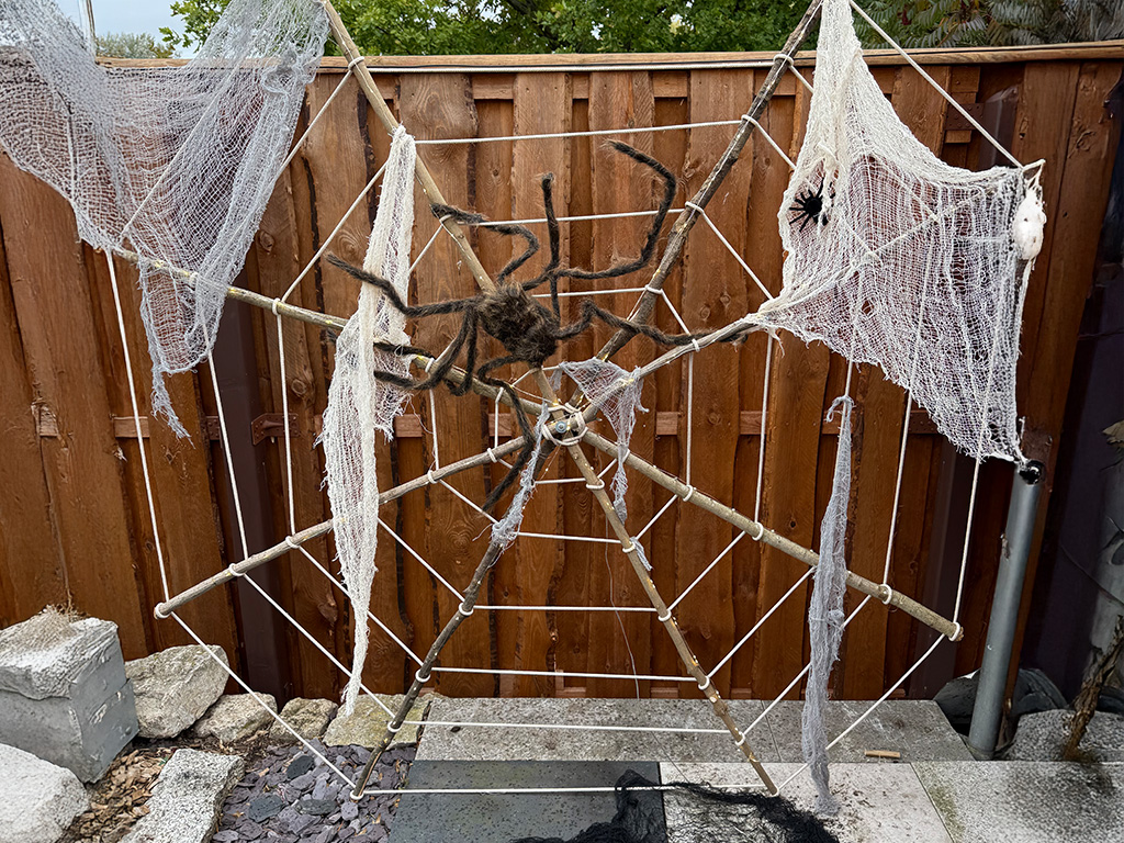 Großes Spinnennetz aus Seilen mit weißen Tüchern und zwei künstlichen Spinnen, auf Holzrahmen im Garten montiert.