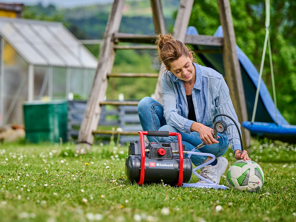 A woman uses a portable Einhell compressor in the garden to inflate a soccer ball.