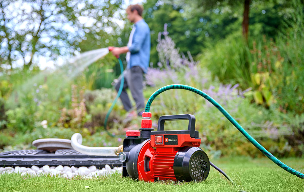 Ein Mann bewässert den Garten, während eine Gartenpumpe im Vordergrund sichtbar ist.