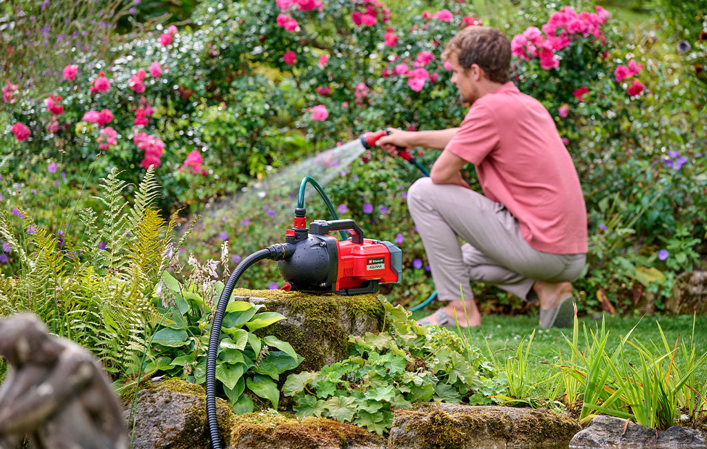 Ein Mann kniet im Garten und nutzt eine Einhell Gartenpumpe zum Bewässern der Pflanzen.