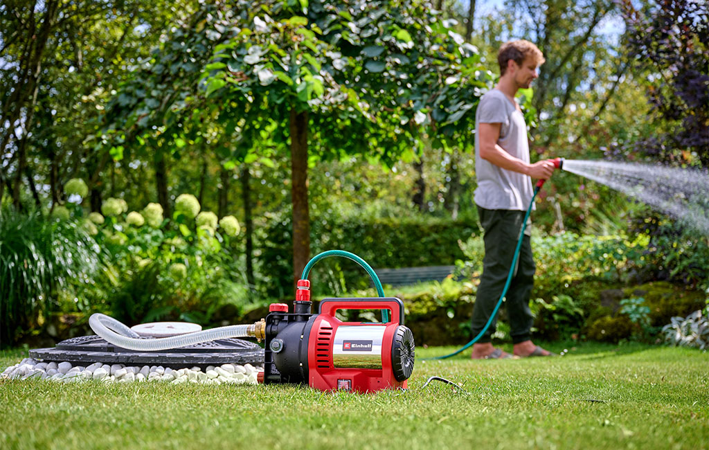 Ein Mann bewässert den Garten mit einer roten Einhell Gartenpumpe im Vordergrund.