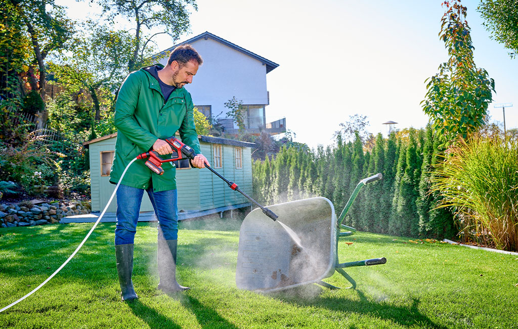 A man in a green jacket and rubber boots is cleaning an overturned wheelbarrow with a cordless pressure washer in a well-kept garden.