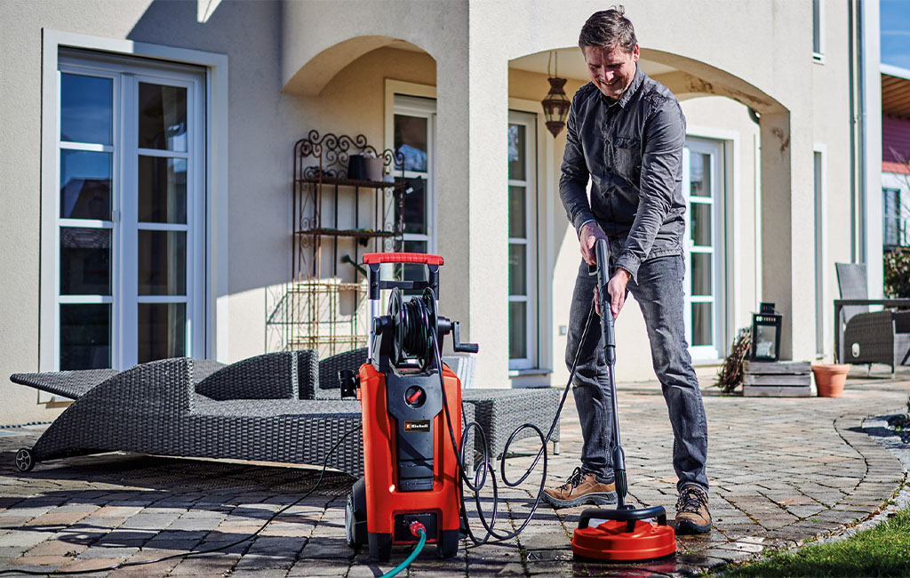 A man is cleaning a paved terrace in front of a house with an Einhell high-pressure washer, with a lounge chair visible in the background.