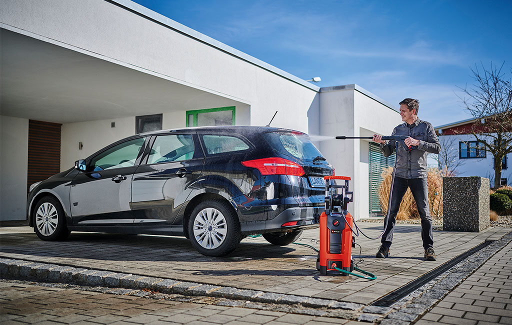 A man is washing a black car with an Einhell high-pressure washer in front of a modern house.