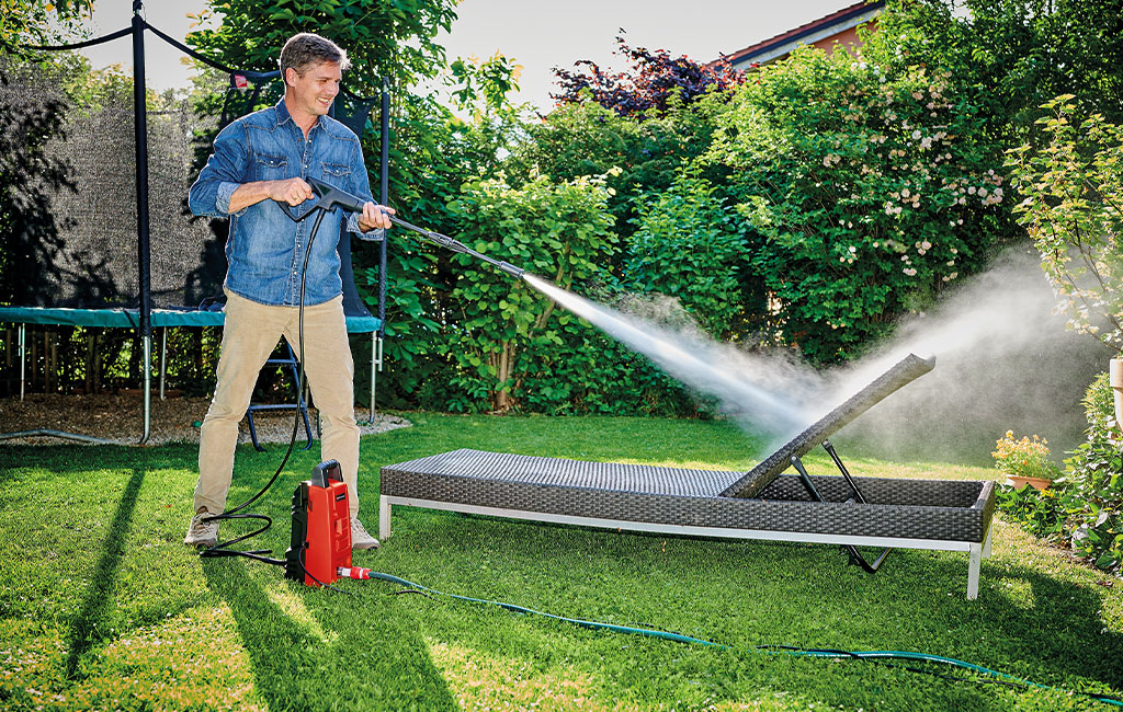 A man is cleaning a garden lounger with a red Einhell high-pressure washer on a lawn in a sunny garden.