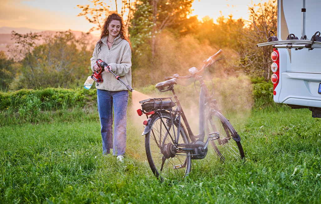 Eine Frau reinigt bei Sonnenuntergang mit einem Hochdruckreiniger ein Fahrrad neben einem Wohnmobil auf einer Wiese.