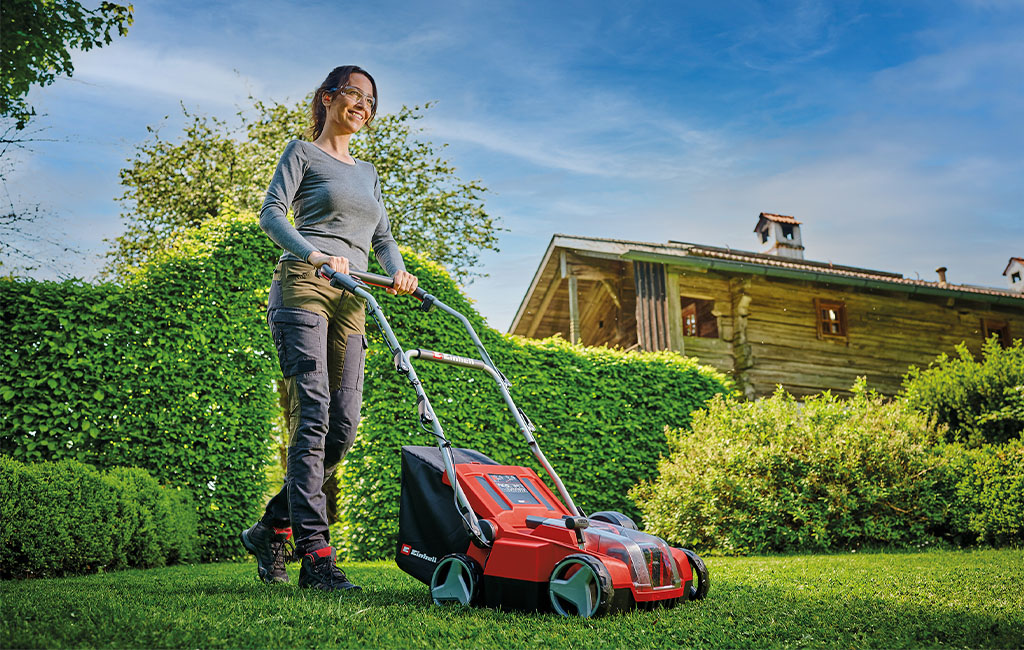 A woman aerating the lawn with a cordless scarifier aerator in the garden in front of a wooden house.