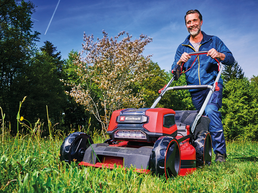 A man is mowing a meadow with a Einhell cordless lawnmower in a green, tree-rich environment on a sunny day.