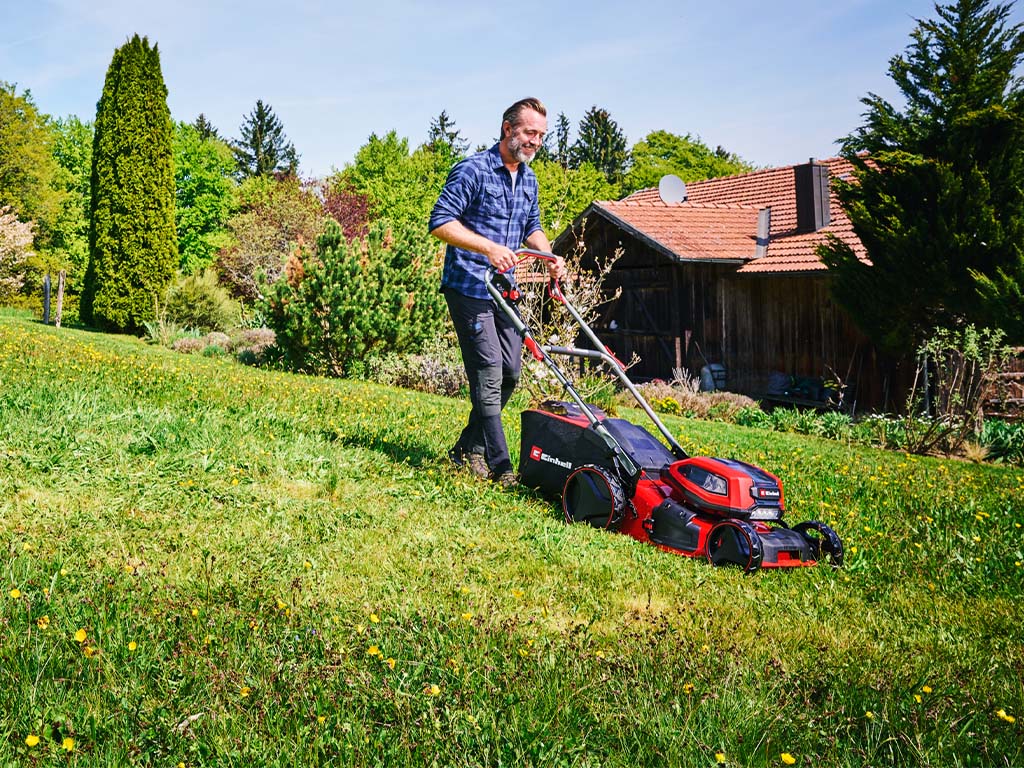 A man is mowing a blooming meadow with a cordless lawnmower in front of a house.