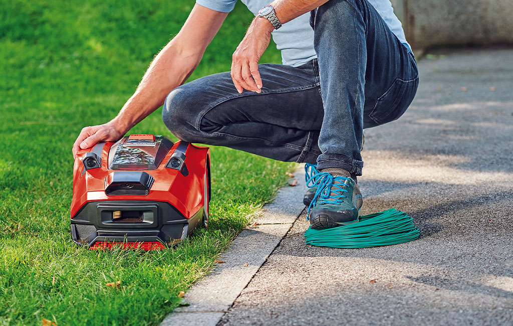 A man kneeling on the ground next to a robotic lawnmower and a boundary wire