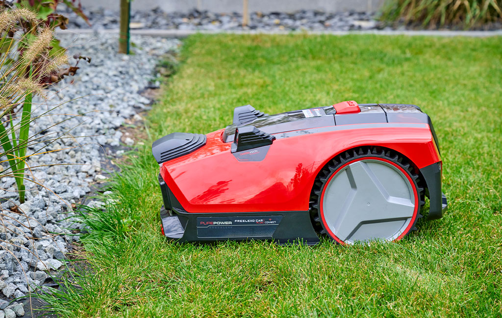 An Einhell FREELEXO CAM robotic lawnmower on a green lawn in front of a stony border