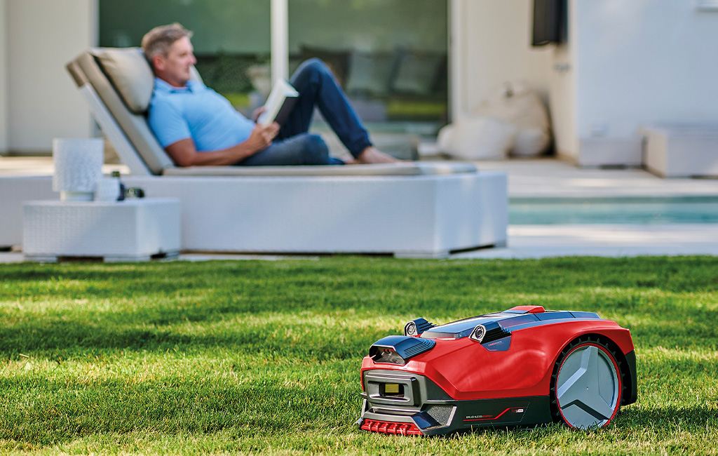 Einhell Robot lawn mower FREELEXO CAM is mowing the lawn while a man relaxes on a lounger