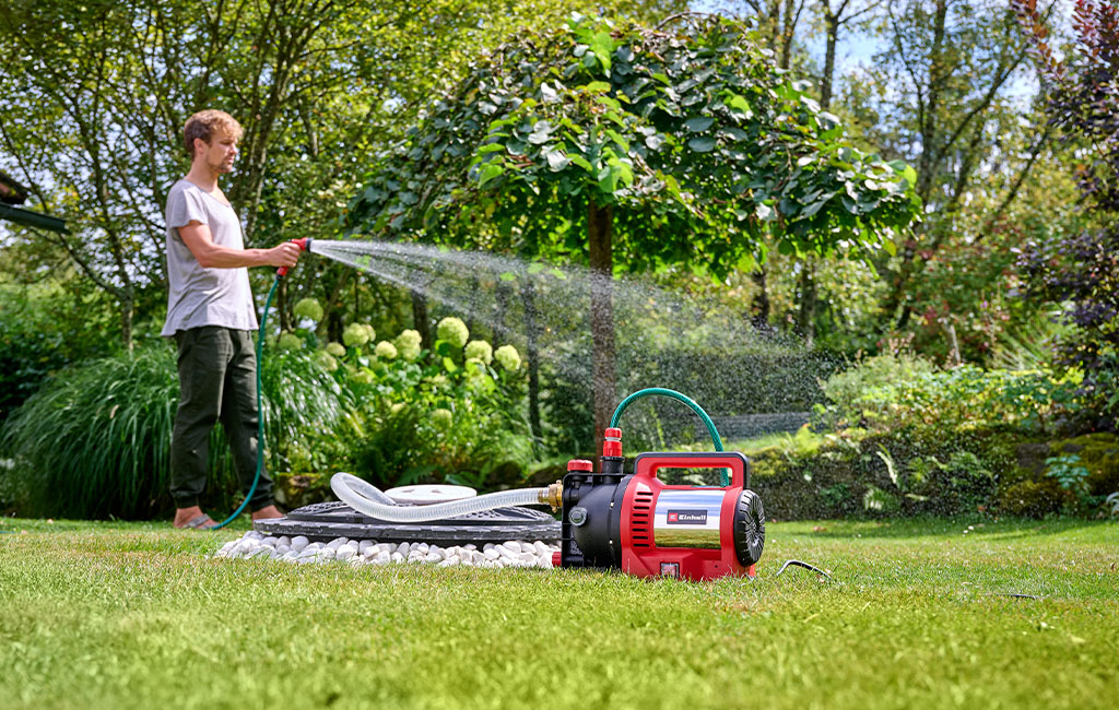 A man waters the garden barefoot using an Einhell garden pump placed on the lawn.