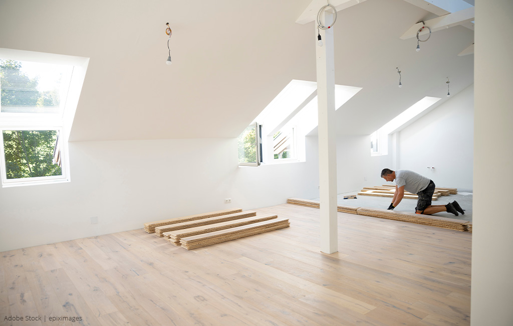 A person is laying laminate flooring in a bright attic room.