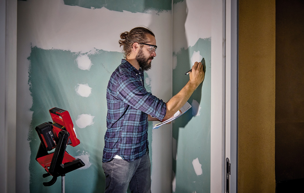 Ein Mann zeichnet mit Bleistift an eine Trockenbauwand, die von einer Akku-Lampe angeleuchtet wird.