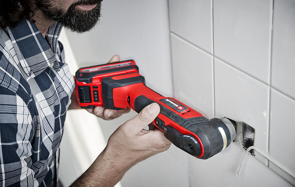 A man uses an Einhell multi-tool to remove a silicone joint from tiles on the wall.