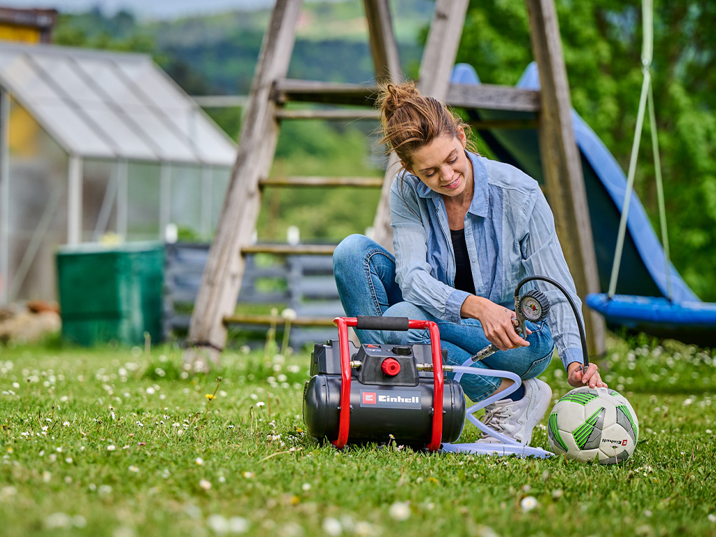 A woman uses a portable Einhell compressor in the garden to inflate a soccer ball.