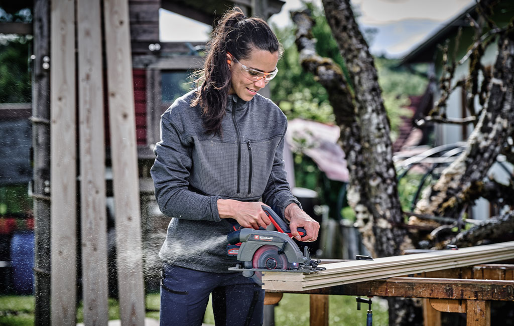 Eine Frau mit Schutzbrille sägt Holz mit einer Handkreissäge im Freien.