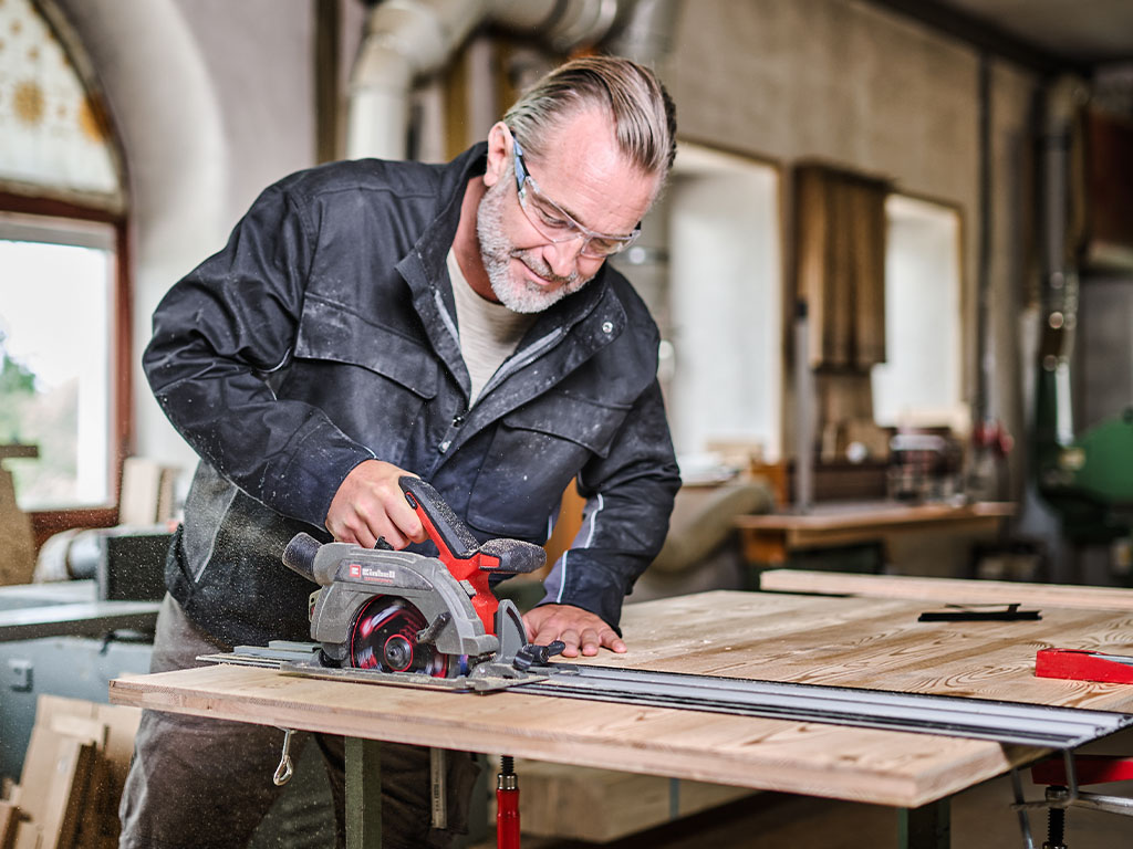 An older man wearing safety glasses is working with a handheld circular saw from Einhell in a workshop.