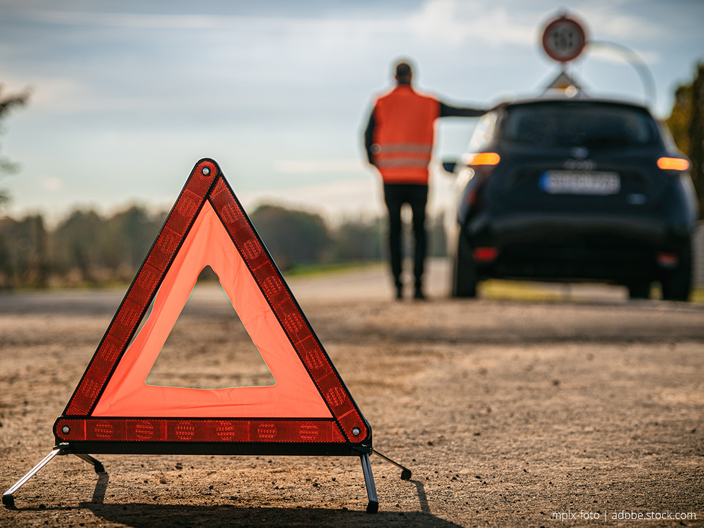 Man in orange safety vest stands leaning against his broken-down vehicle and an orange warning triangle can be seen in the foreground.