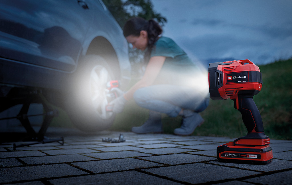 A woman uses a portable work light to change a car tire at night.