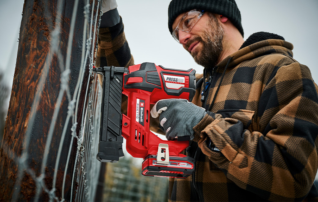 A man secures wire to a fence using an cordless stapler in an outdoor setting.