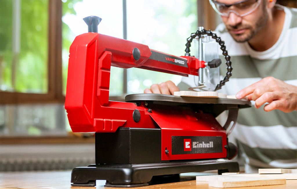 A man works on a piece of wood using a red Einhell scroll saw on a table.
