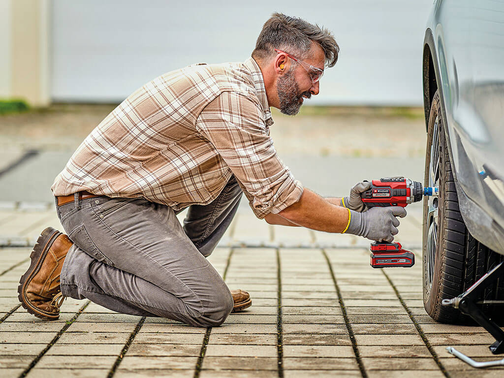 A man kneels on the ground using the Einhell cordless impact wrench to tighten a wheel nut on a jacked-up car.