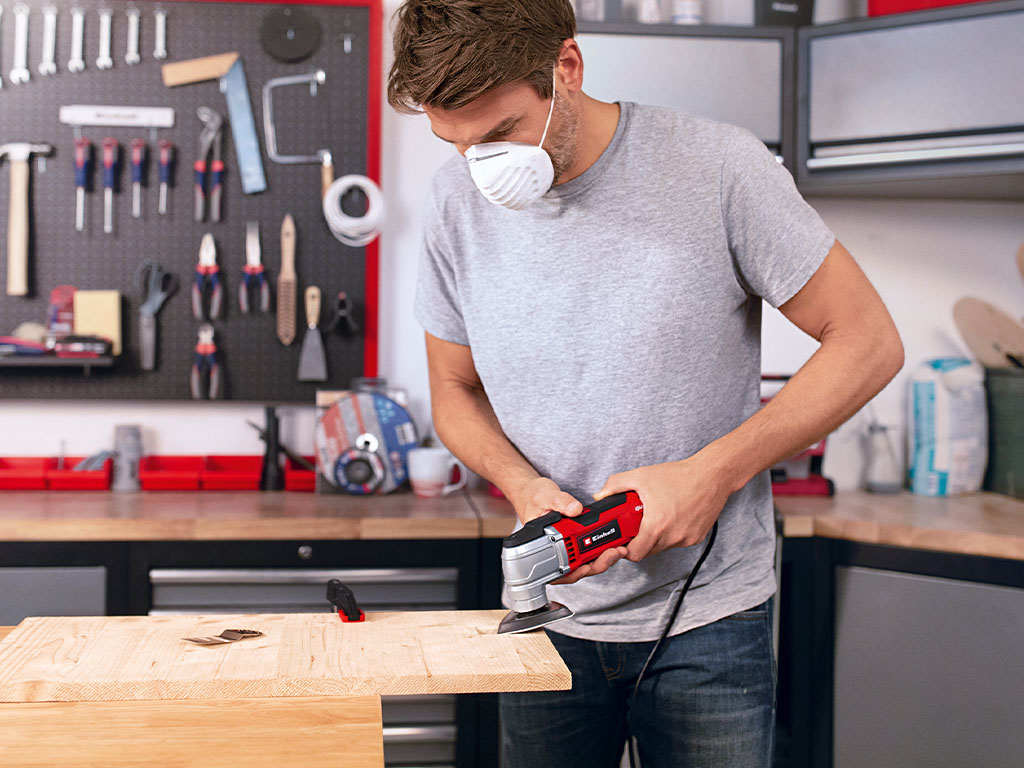 A man holds a red multifunctional tool against a wooden beam, precisely working on the surface.