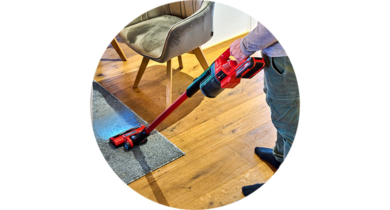 A person vacuums a carpeted floor with a cordless Einhell vacuum cleaner on wooden flooring.