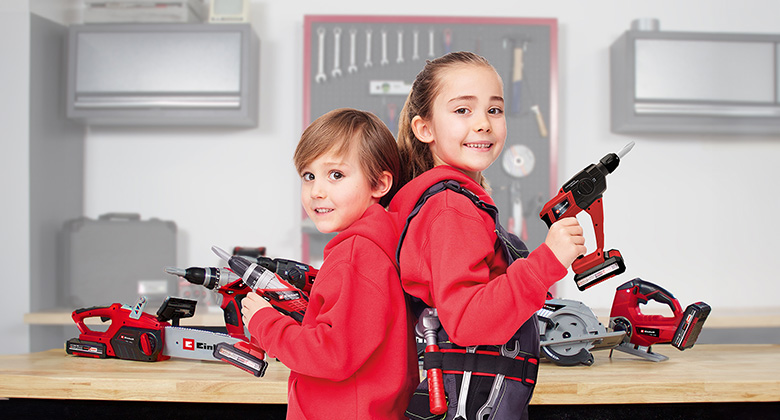 Two children holding Einhell kids' toy tools stand back-to-back in a workshop.