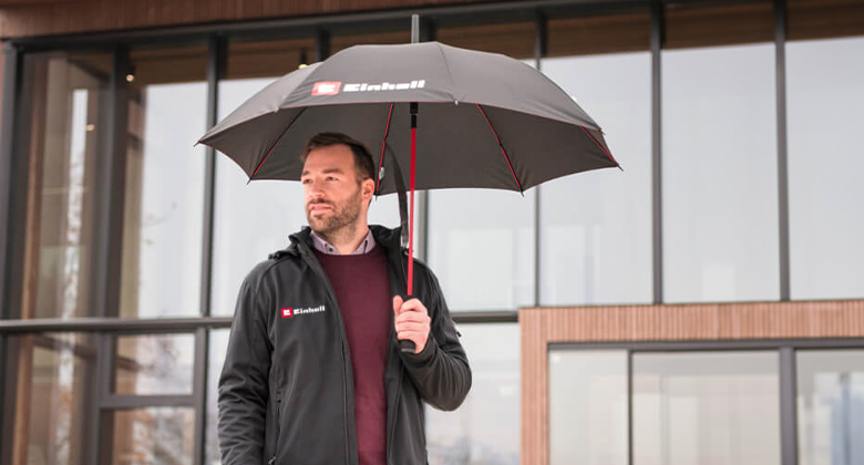 A man wearing an Einhell jacket holds an Einhell umbrella in front of a modern building.