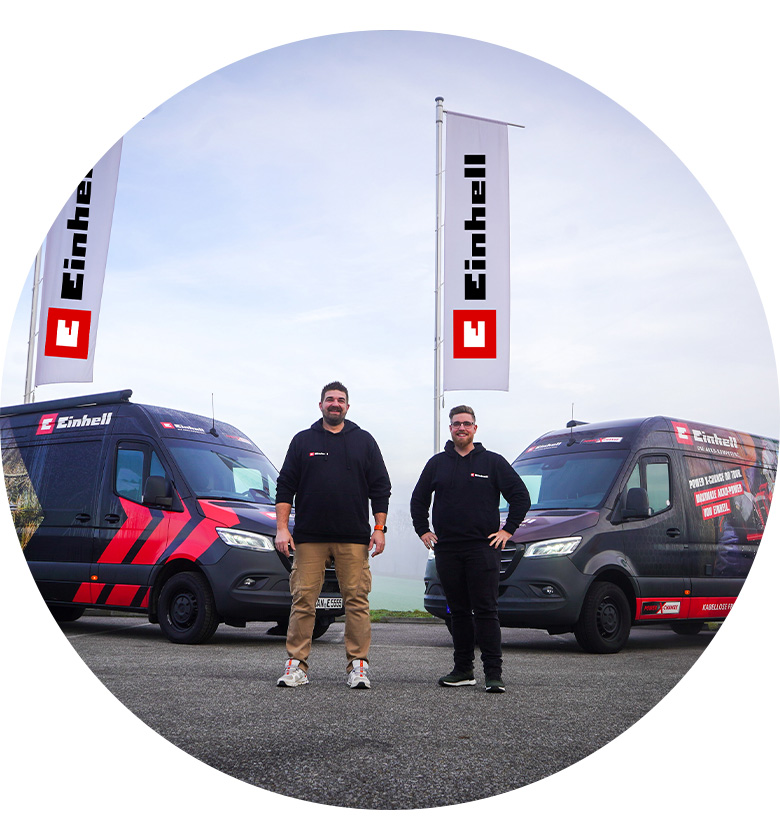 Two men in black hoodies stand in front of two Einhell vans with Einhell flags in the background.