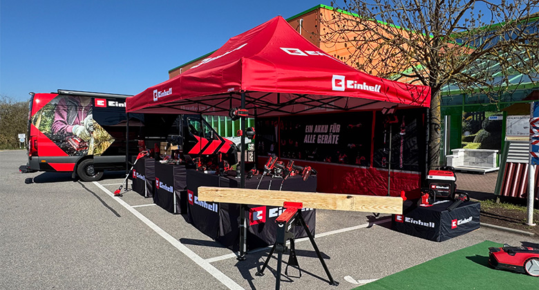 Ein rotes Einhell-Promotionzelt mit dem Slogan „Ein Akku für alle Geräte“ auf einem Parkplatz, umgeben von Werkzeugen, Geräten und einem Einhell-Transporter.