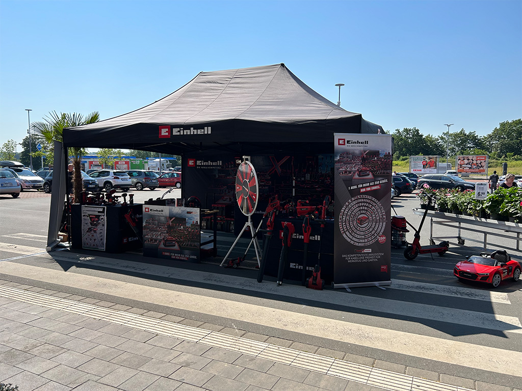 Outdoor Einhell promotional tent with a spinning wheel and product displays, set in a parking lot.