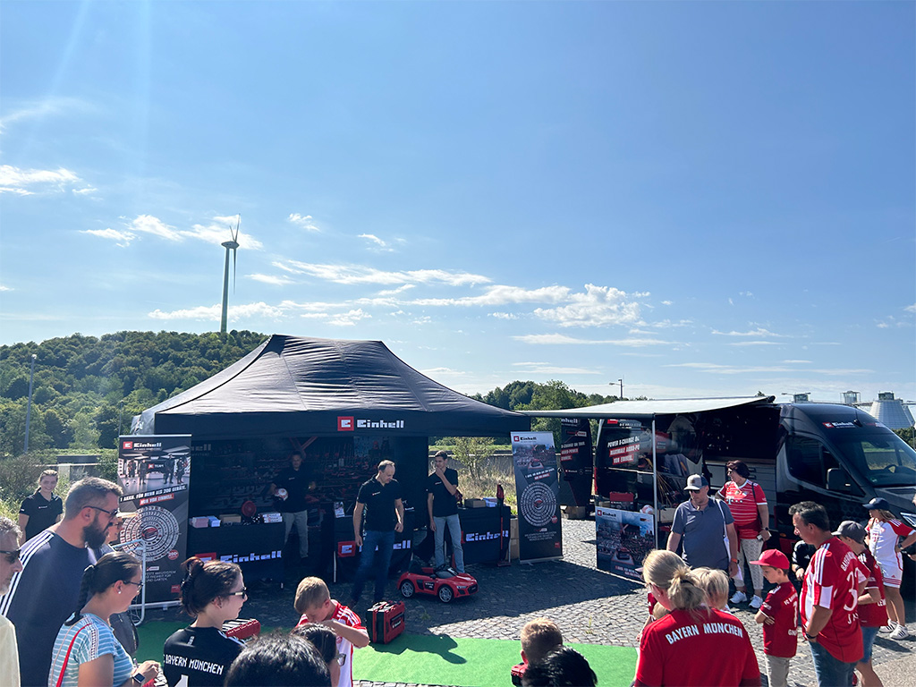 Outdoor Einhell promotional booth with visitors exploring tools and products, featuring a branded van and banners in a sunny setting.