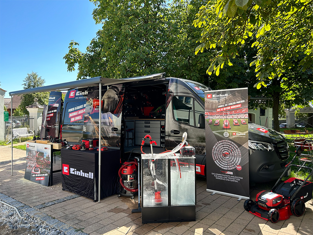 Einhell van and booth with displayed tools and promotional banners under the shade of trees.