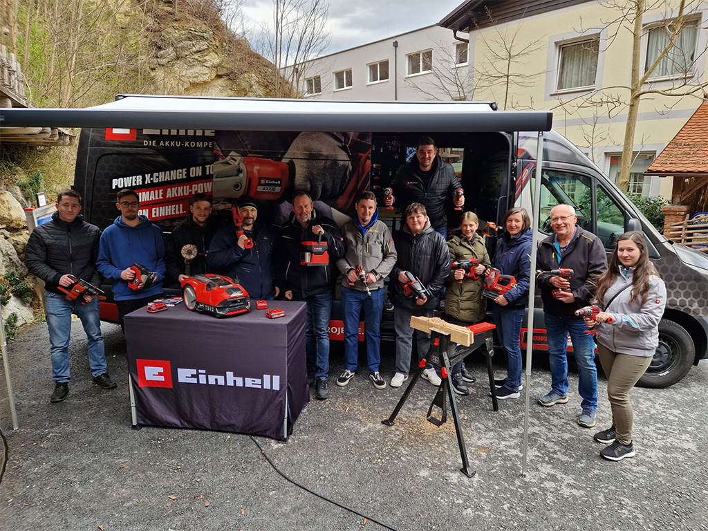 Group photo of visitors and Einhell team members holding tools in front of the promotional van outdoors.