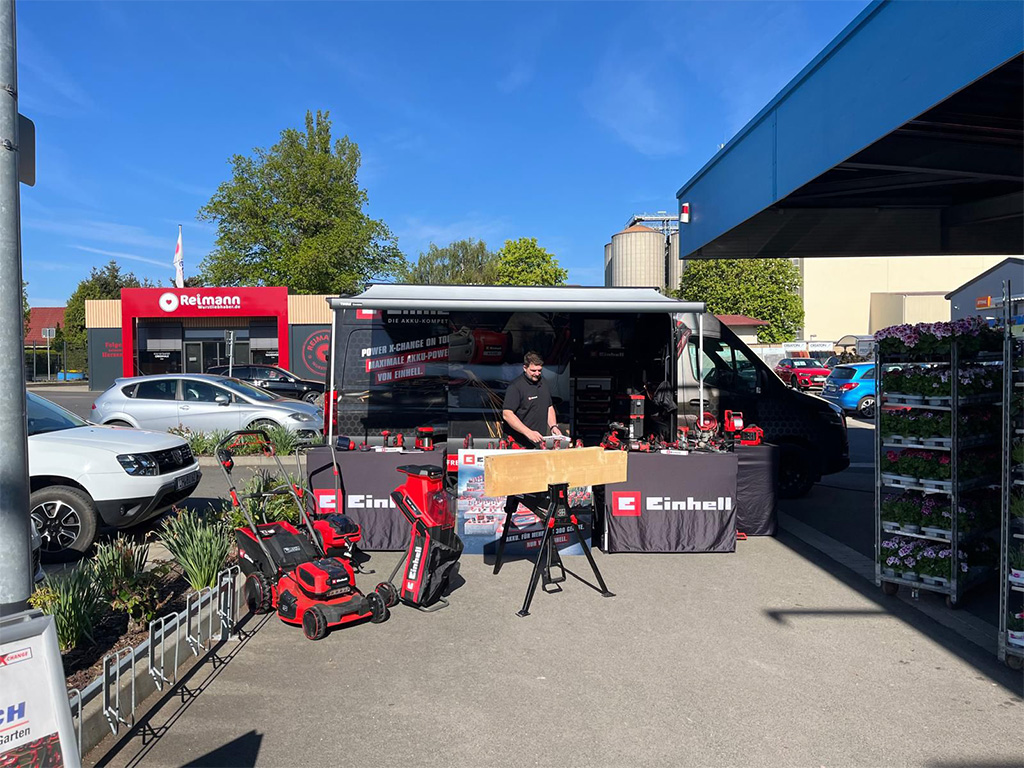 A sunny outdoor Einhell promotional setup showcasing tools and lawn equipment in front of a retail store.