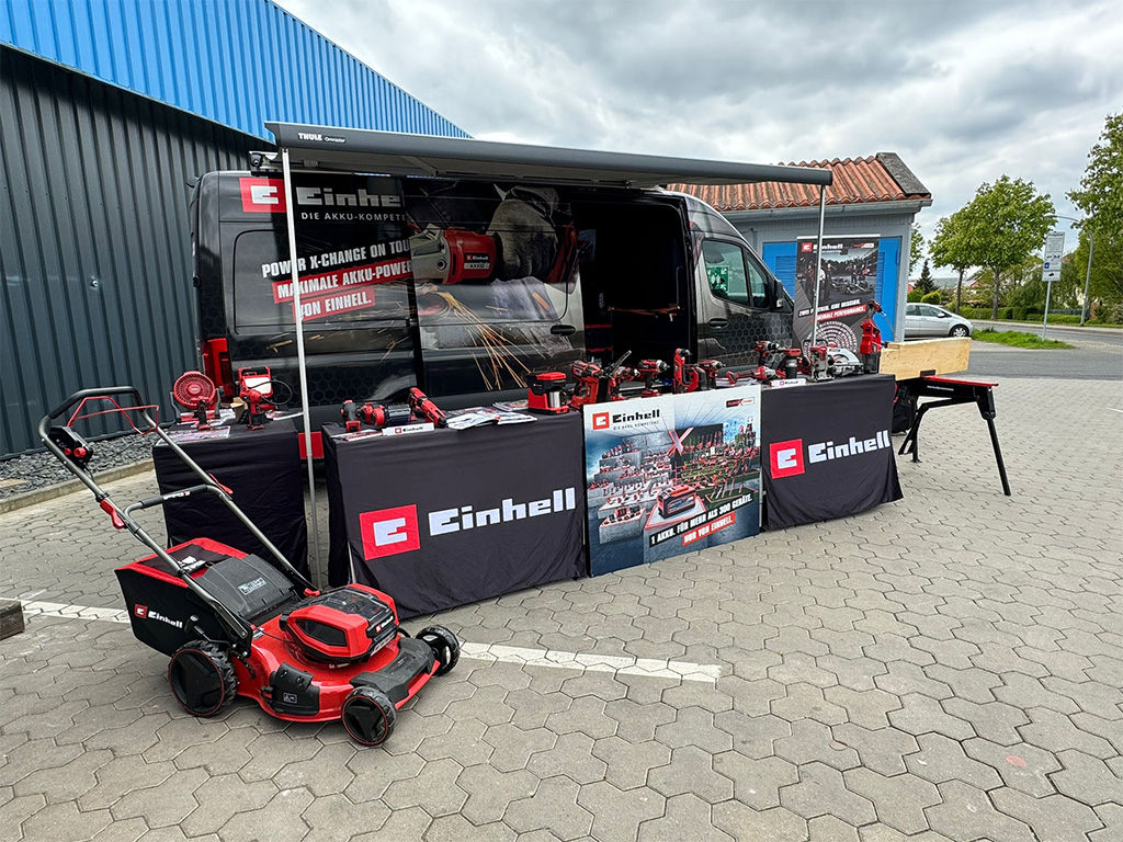 Einhell promotional van and booth displaying tools and garden equipment on a paved surface near a retail building.